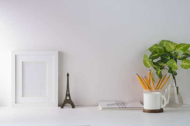 Front view blank picture frame, houseplant, pencil holder, book and coffee cup on white table. Minimal workplace, Copy space for your text. - 写真・画像