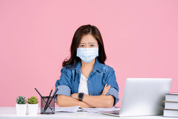 若いアジアのビジネスマン女性でカジュアル服とマスクを身に着けていることを防ぐためにcovid 19座って仕事でオフィスデスクでPCラップトップで隔離されたピンクの背景スタジオ. - 写真・画像