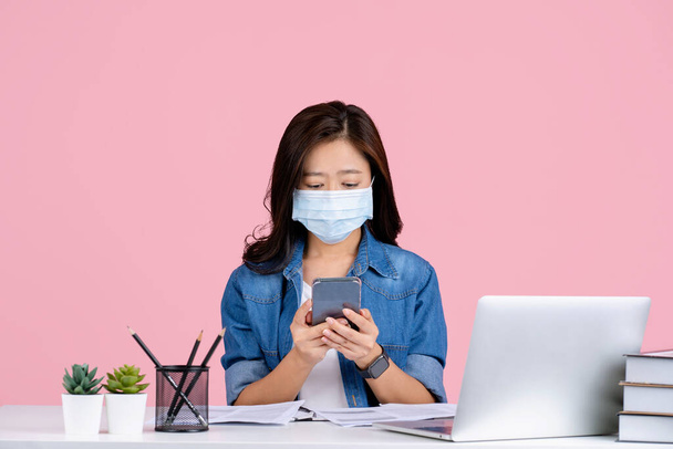 Jonge Aziatische zakenvrouw in casual kleding en het dragen van een masker om te voorkomen dat covid 19 zitten werk op kantoor bureau met pc laptop geïsoleerd op roze achtergrond studio. Ze gebruikt een smartphone. - Foto, afbeelding