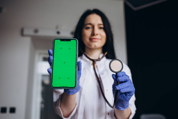 Крупним планом усміхнена дружня молода жінка-лікар кардіолог тримає мобільний телефон з макетним екраном і стетоскопом, рекламуючи мобільні програми відстеження здоров'я, концепцію телемедицини
. - Фото, зображення