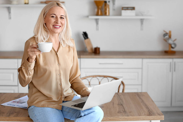 Утро зрелой женщины с ноутбуком, пьющей кофе на кухне - Фото, изображение