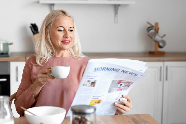 Morning of mature woman drinking coffee and reading newspaper in kitchen - Photo, Image