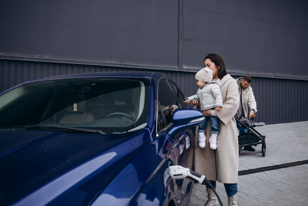 Mother and daughter charging battery of electric car. In the background, Dad takes the stroller out of the trunk - Фото, изображение