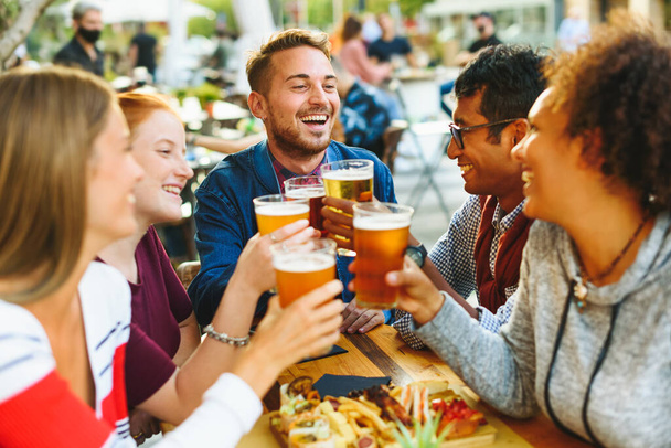 Groep van opgetogen multiraciale vrienden die elkaar aankijken terwijl ze juichen met bierglazen aan tafel met eten op het terras van de pub - Foto, afbeelding