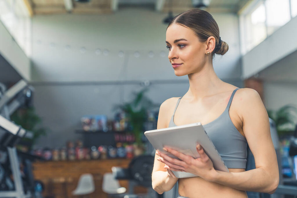 Indoor portrait of a young white millennial woman with slicked back hair and gray sports bra holding a tablet, creating workout program for her clients. High quality photo - Foto, afbeelding