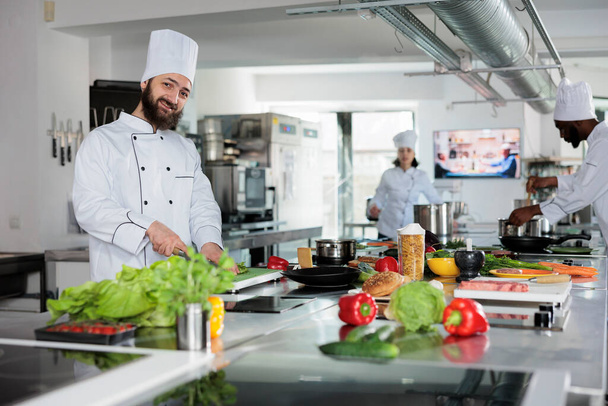 Gurmánská kuchyně kavkazská kuchařka připravuje recepty na jídlo a zároveň krájí čerstvou ekologickou zeleninu. Usmívající se hlava kuchař vaření gastronomické jídlo při přípravě zdobení. - Fotografie, Obrázek