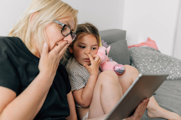 Granny and preschool girl using tablet together at home on the sofa, family togetherness time . High quality photo - Fotó, kép