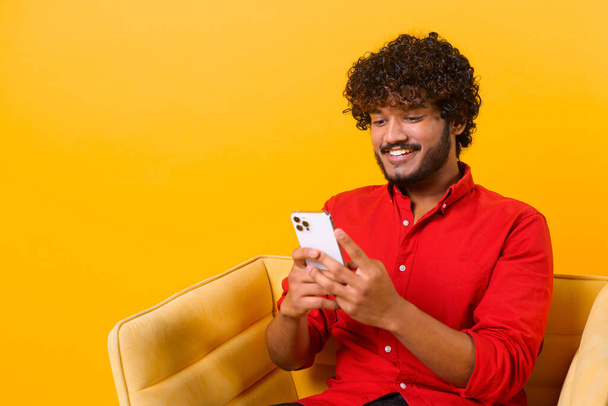 Waist up view of happy handsome blogger young adult man sitting at the armchair, using smartphone and texting message with smile. Indoor, isolated, studio shot, orange background - Фото, зображення