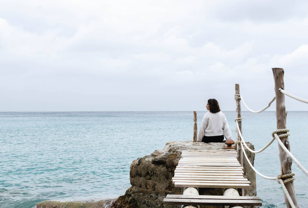 mujer joven admirando el paisaje de IBIZA. sonríe mirando a la izquierda. chica sentada en una roca en el mar Mediterráneo. cruzó un viejo puente de madera en malas condiciones - Foto, Imagen
