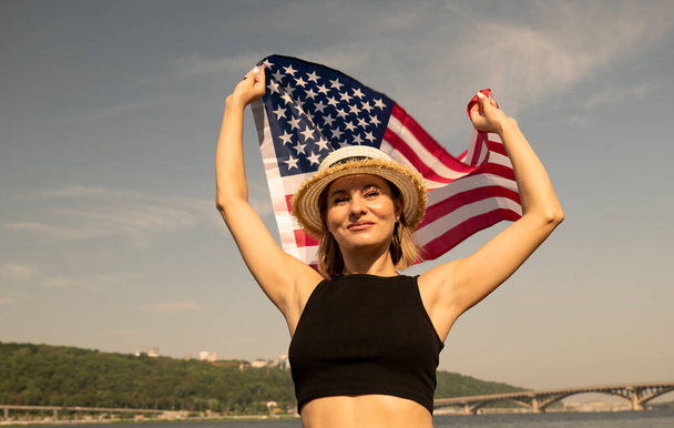 Widok z dołu młodej kobiety w kapeluszu z flagą USA w pobliżu rzeki w mieście. Dzień Niepodległości 4 lipca. wakacje w Ameryce - Zdjęcie, obraz