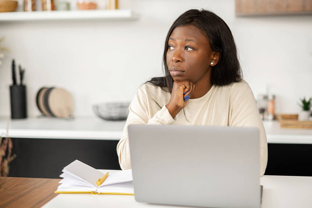 Worried African-American woman sits with a laptop in the kitchen at home, sad because of received rejection, tired from online work, young female feels depressed - Photo, image