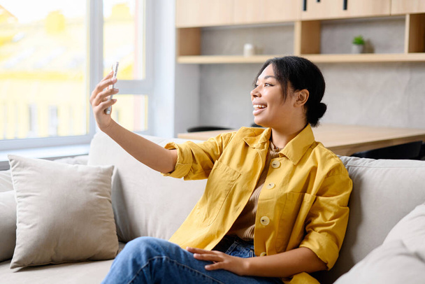 Positive young woman using the smartphone for making selfie. Smiling female has pleasant online meeting or shooting herself while sitting on the couch at home - Photo, image