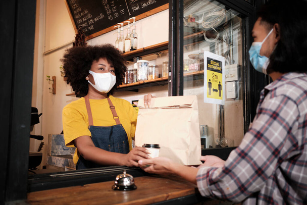 Africká americká baristka s obličejovou maskou pracuje v kavárně se společenskou vzdáleností, donáška kávy pro zákazníka, nová běžná obsluha malé kavárny v karanténním životním stylu COVID19. - Fotografie, Obrázek