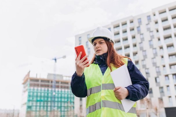 Architect at a construction site. Portrait of woman constructor wearing white helmet and safety yellow vest. Upset, skeptical, serious woman looking at the phone outdoors. - Photo, Image