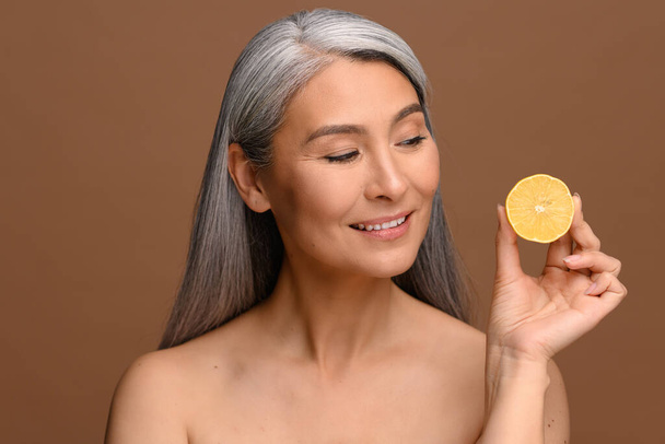 Headshot of charming middle-aged gray-haired Asian woman holding half of lemon isolated on bronze background, mature lady with naked shoulders standing with piece citrus and looking at it - Fotó, kép