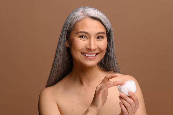 Enchanting topless middle aged Asian woman looks at the camera and laughing happily, holding jar of moisturizer cream isolated on brown background, charming korean lady advertising cosmetology product - Fotó, kép