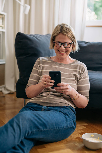 middle adult woman sitting on the floor and using mobile phone at home - Foto, Imagem