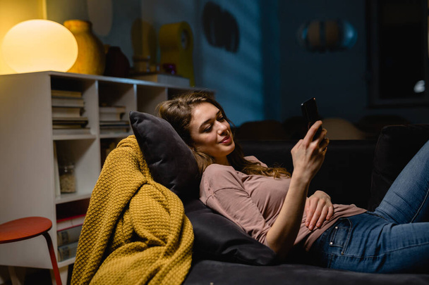 woman relaxing on sofa and texting messages on smartphone. evening night scene - Фото, зображення