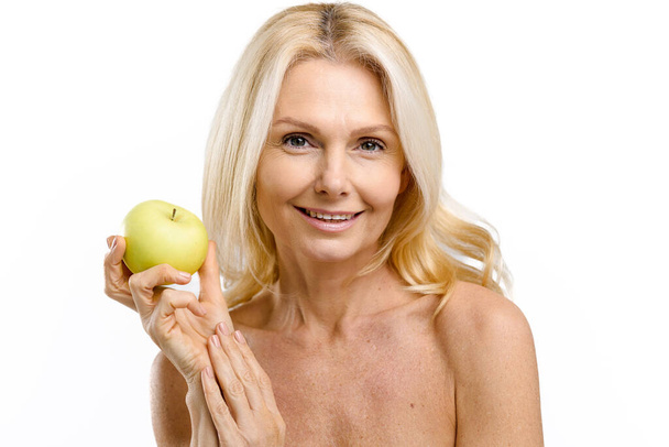 Middle-aged model with naked shoulders isolated on white, demonstrates smooth and radiant skin. Cheerful joyful mature 50s woman posing with fresh green apple and smiling. Anti age treatment concept - Fotó, kép
