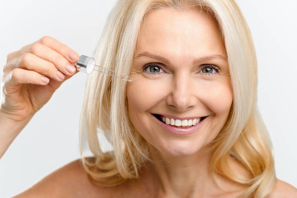 Close-up portrait of magnificent middle-aged blonde woman using skin care product, charming lady with radiant smooth face skin pipette of anti-aging serum smiling isolated on white - 写真・画像
