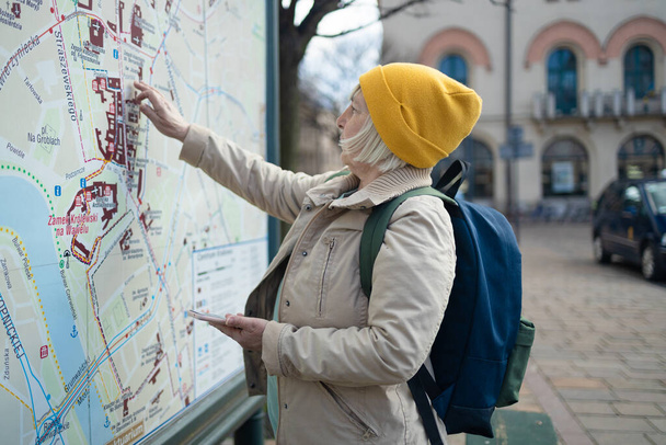 Блондинка 50-х годов туристка с рюкзаком прокладывает маршрут на карте стенда в городе. Концепция путешествия и туризма. - Фото, изображение