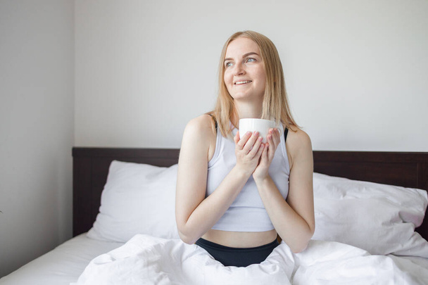Cheerful 30a European female in white sleepwear sitting with cup of coffee on bed and having breakfast while enjoying morning in bedroom at home.  - Fotoğraf, Görsel