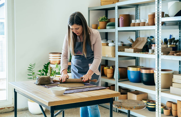 Small Business: Lachende vrouw pottenbakker met behulp van deegroller voor klei massa en het maken van keramiek object op een bureau in haar studio - Foto, afbeelding