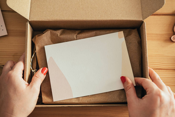 Table Top View Of An Anonymous Woman Packing Gift Tag On A Merchandise For Delivery At Her Store. Small Zero Waste Business: Close up photo of woman hands putting blank thank you note in a cardboard box on a wooden desk in her workshop. - Photo, Image