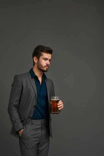 Concentrated caucasian brewer entrepreneur with hand in pocket holding glass of beer. Bearded man wearing suit. Brewing business. Beer tasting. Isolated on grey background. Studio shoot. Copy space - Fotografie, Obrázek