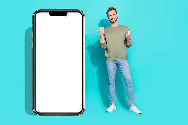 Full size photo of young guy index promo hold telephone wear shirt jeans footwear isolated on teal background. - Foto, imagen