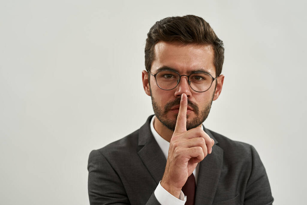 Portrait of caucasian businessman showing silence gesture. Bearded man wearing suit and glasses. Concept of modern successful male lifestyle. Isolated on white background. Studio shoot. Copy space - Φωτογραφία, εικόνα
