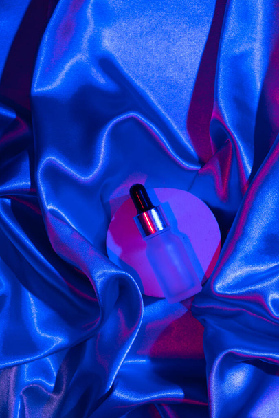 Csepegtető üveg kozmetikai olajjal vagy szérummal textilen neon fényben. Trendi színek és fények. - Fotó, kép