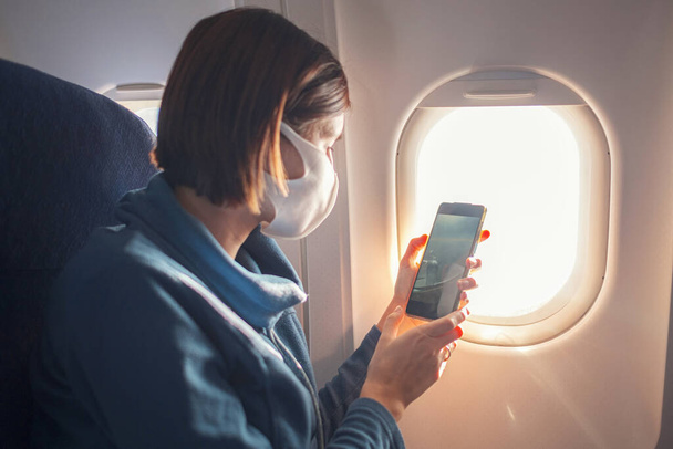 Young beautiful woman sitting at window of plane during the flight. new normal travel after covid-19 pandemic concept. taking photo from an airplane window - 写真・画像
