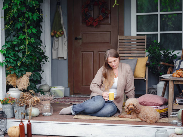 femme caucasienne d'âge moyen en cardigan brun assis sur la terrasse de la maison avec son caniche beige et profiter du temps chaud d'automne et boire le café du matin, concept de style de vie d'automne - Photo, image