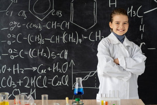 Smiling little student scientist making experiments in laboratory. Schoolgirl standing near blackboard with chemical formulas. Chemistry in school. Early development of children. Educational concept. - Foto, Imagen