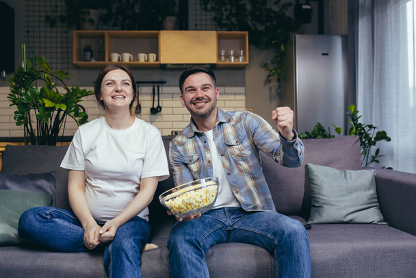 Married couple man and pregnant woman together on sofa together watching sports match cheering and eating popcorn - Foto, imagen