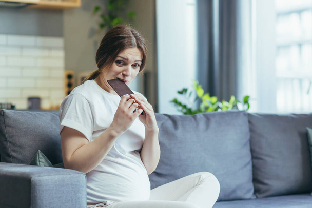 Young pregnant woman at home sitting on the couch eating chocolate - Φωτογραφία, εικόνα