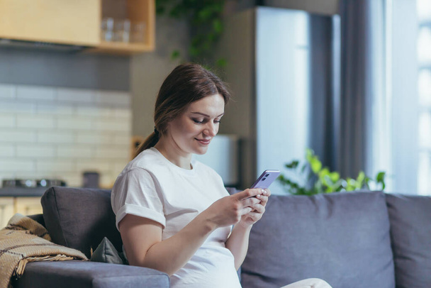 Pregnant woman at home uses the phone while sitting on the couch - Fotoğraf, Görsel