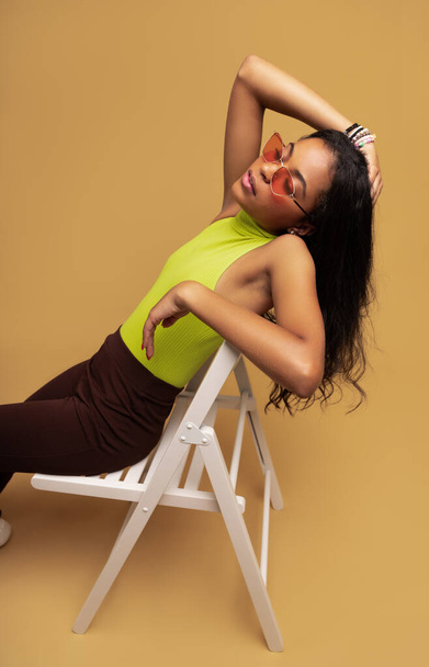 Jeune femme hispanique à la mode en vêtements décontractés et lunettes de soleil touchant de longs cheveux noirs tout en se reposant sur une chaise sur fond ocre - Photo, image