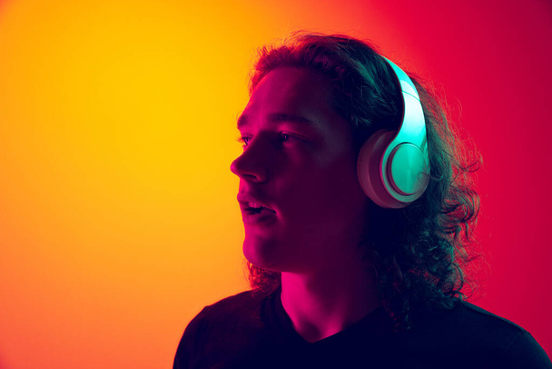 Young excited man with long curly hair listening to music in headhones isolated on gradient yellow-pink background in neon. Concept of beauty, fashion, youth culture and emotions. Male portrait. - 写真・画像