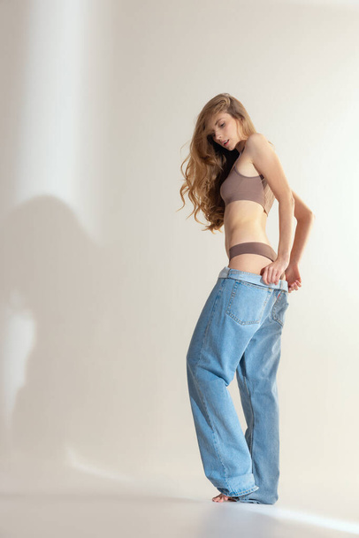 Full-length portrait of slim beautiful woman in underwear putting on oversized jeans isolated over grey studio background. Weight-loss. Concept of beauty, body and skin care, femininity, ad - Foto, Bild