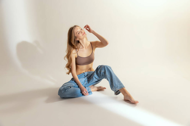 Портрет молодої стрункої жінки в бежевому верхньому і синьому джинсах, що сидять на підлозі, позує ізольовано на сірому студійному фоні. Самолюбство. Поняття краси, догляду за тілом і шкірою, мода, стиль, жіночність, реклама
 - Фото, зображення