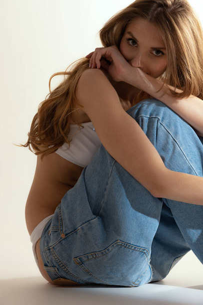 Portrait of tender young girl sitting, posing isolated over grey background. Model wearing white top and jeans. Concept of beauty, youth, lifestyle, body and skin care, wellness. Copy space for ad - Φωτογραφία, εικόνα