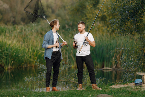 Friends is standing near the river and fishing. Two fishermen talking and drinking a beer. One man wearing white t-shirt and other blue shirt. - Foto, Bild