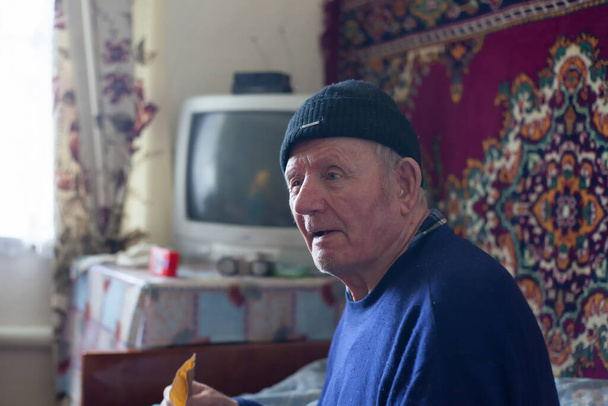 Ein alter Großvater sitzt auf einem Bett in einem alten Zimmer mit einem Fernseher im Hintergrund und einem rustikalen Teppich an der Wand - Foto, Bild