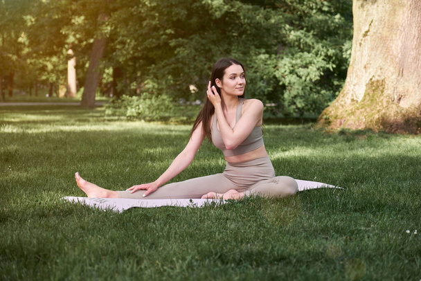 A young woman practices yoga in a city park on a warm summer day. Sports girl sits on the green grass. The concept of a healthy lifestyle. - Photo, Image