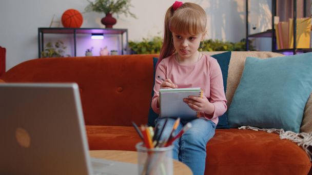 Online onderwijs video oproep op laptop van kleine kind meisje thuis virtuele klasse. Lessen op afstand van het kleine schoolmeisje notities maken, scherm bekijken, tutor luisteren. Modern e-learning op afstand - Foto, afbeelding