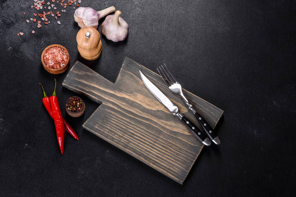 Un plato vacío con un cuchillo, tenedor o cuchara con una tabla de cortar de madera sobre un fondo de hormigón oscuro. Preparación de electrodomésticos e ingredientes para cocinar en casa - Foto, Imagen