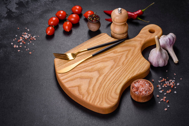 An empty plate with a knife, fork or spoon with a wooden cutting board on a dark concrete background. Preparation of appliances and ingredients for home cooking - Photo, Image