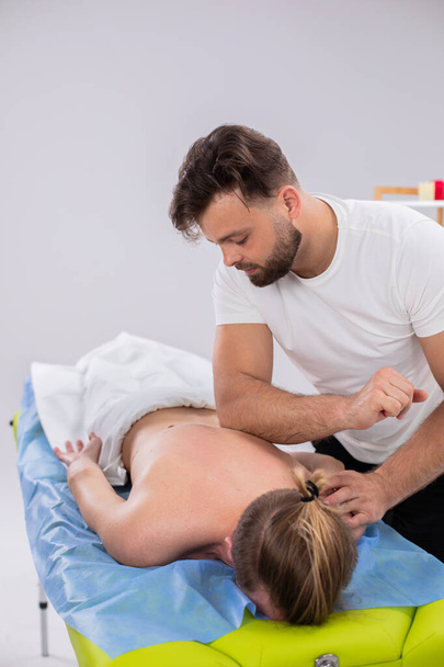 At medical clinic the massage therapist man using a wooden stick to massaging professional the back muscular while the client laying on massage table. - Photo, image